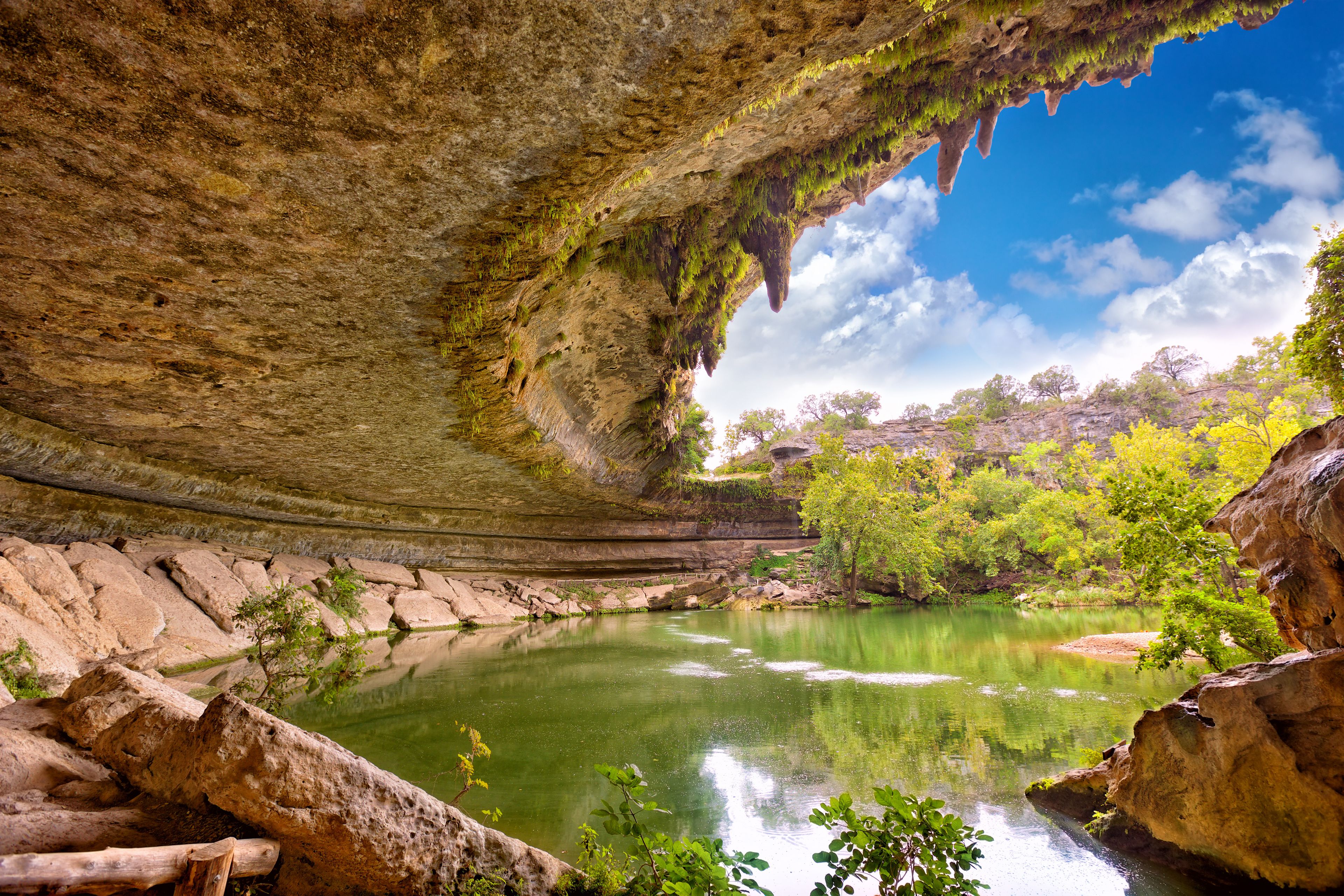 A natural cave opening with a river inside, surrounded by greenery and rocky terrain.