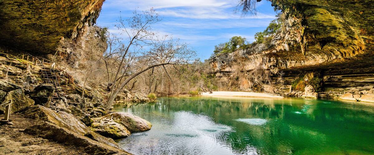 A natural cave opening with a clear blue sky above, a calm turquoise pool, and rocky terrain.