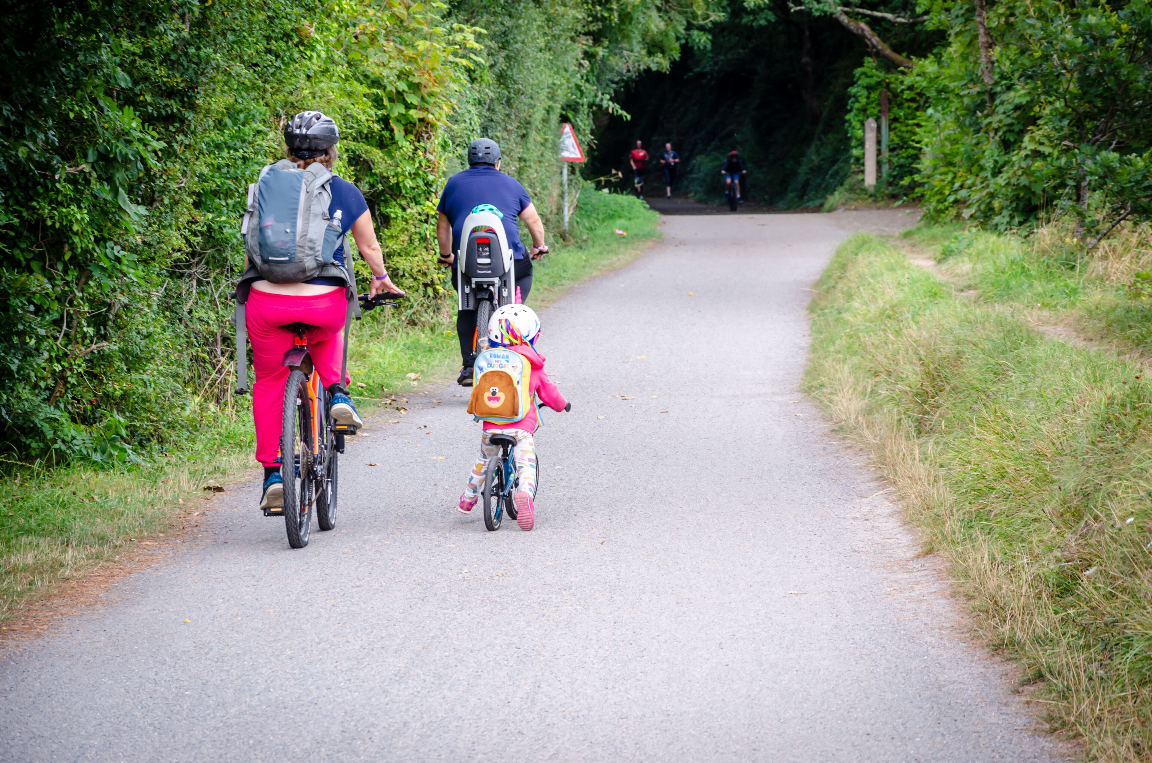 A narrow paved path flanked by greenery, with cyclists and a child on a tricycle.