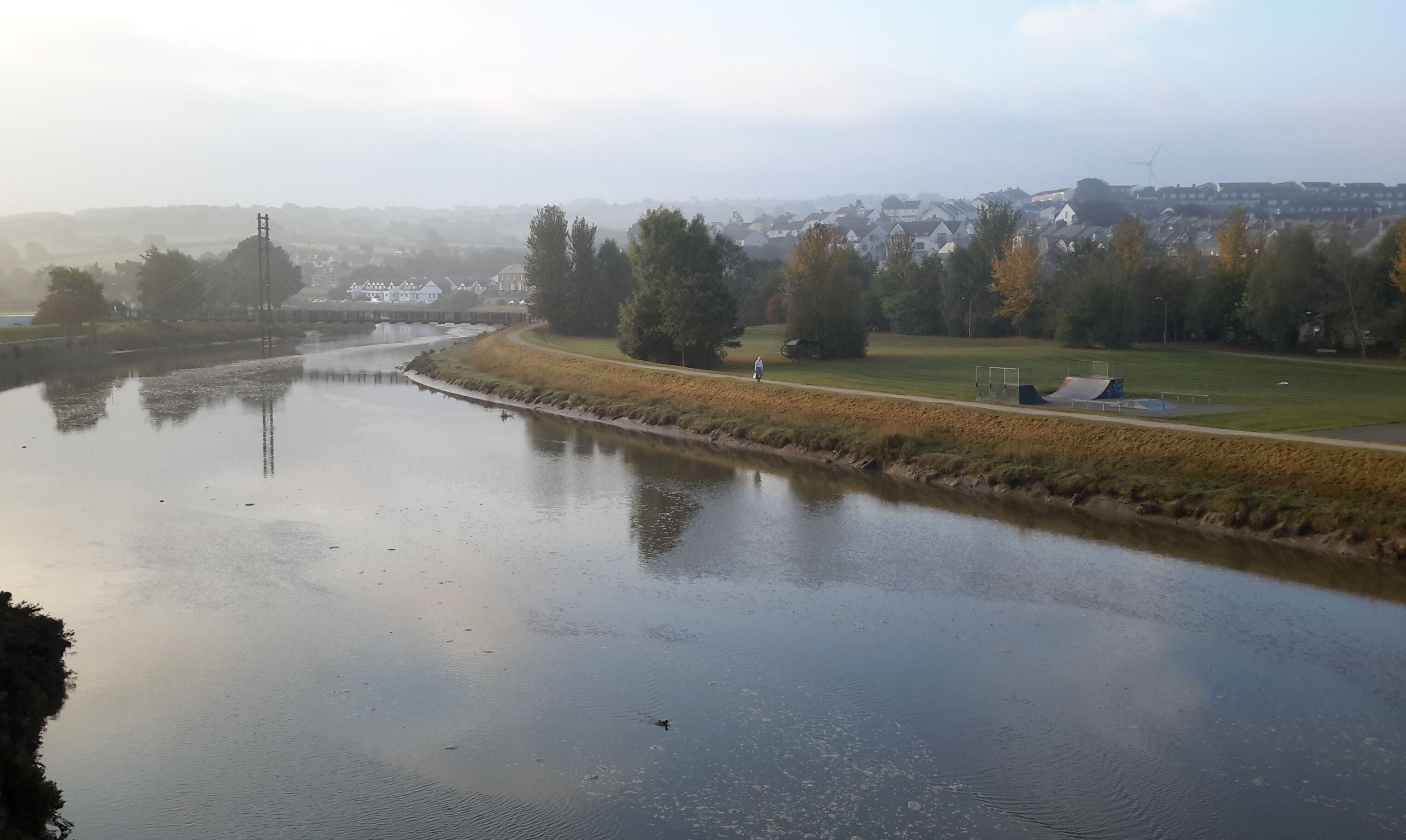 A river with a person walking along the bank, surrounded by trees and a park with sports facilities.