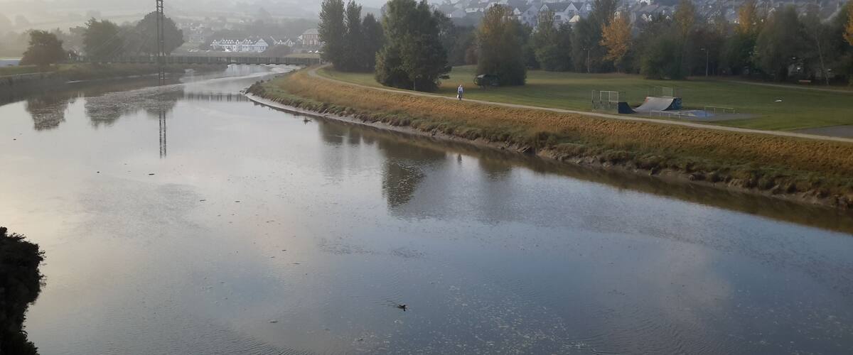 A river with a person walking along the bank, surrounded by trees and a park with sports facilities.
