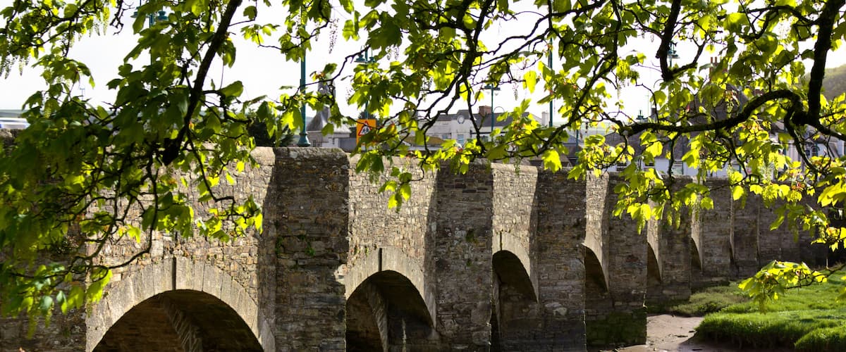 Un puente de piedra con varios arcos sobre un río, rodeado de vegetación y un cielo despejado.