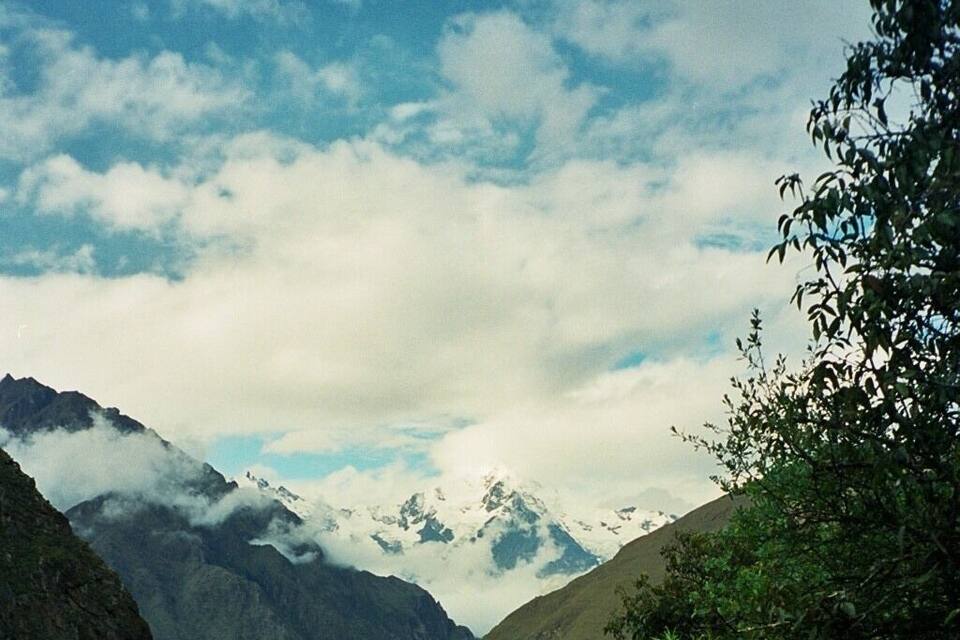 Un sendero montañoso y estrecho rodeado de abundante vegetación y picos nevados bajo un cielo parcialmente nublado.