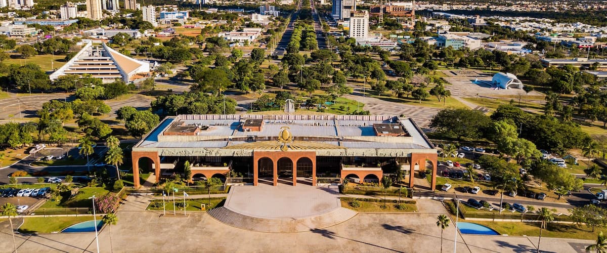 A large, historic building with a central dome and arched entrance, surrounded by a well-maintained park with palm trees and a swimming pool.