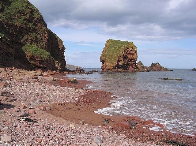 A rocky beach with a prominent rock formation covered in grass, surrounded by cliffs and calm sea.