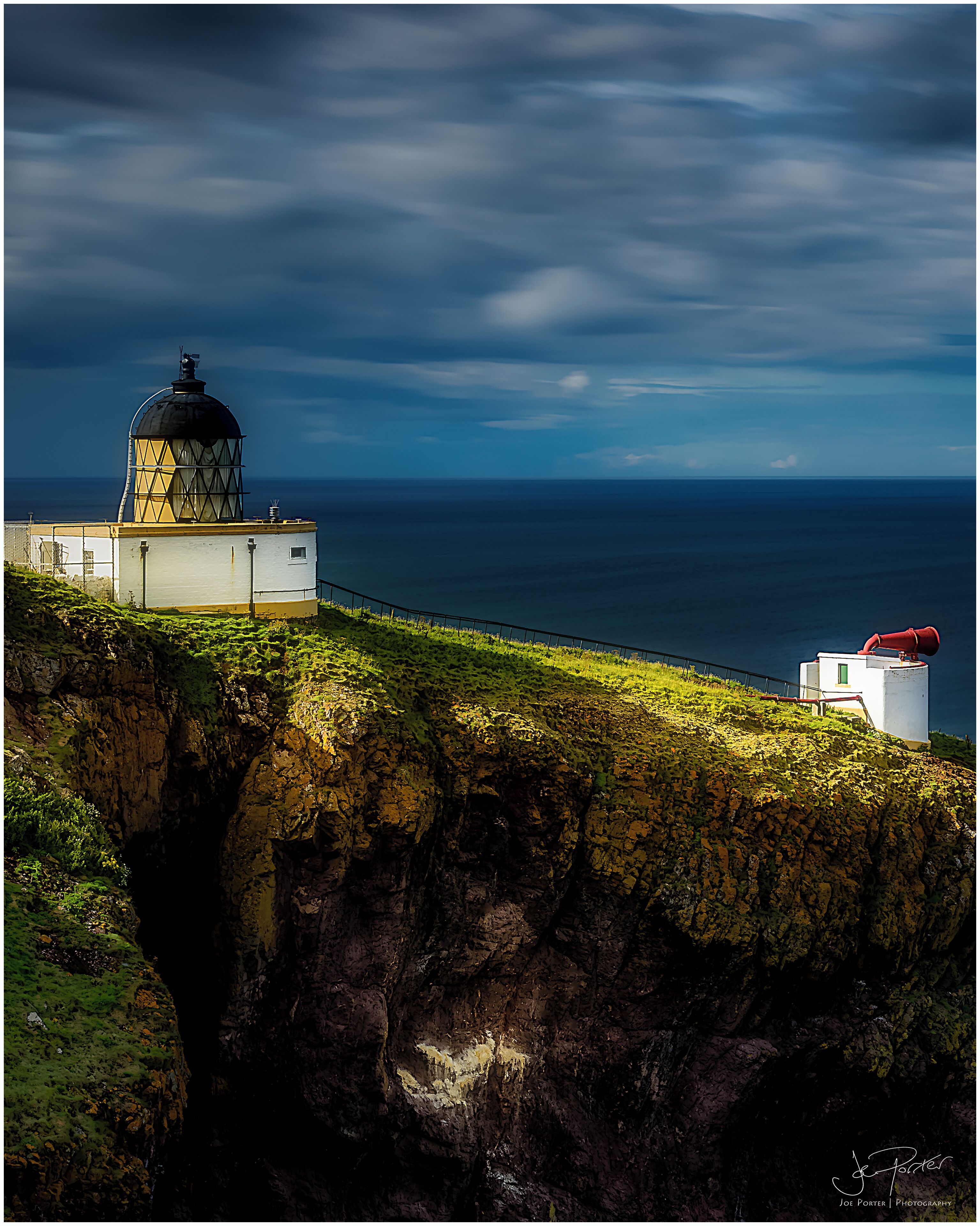 A lighthouse on a cliff overlooking the ocean, with a clear sky and rocky terrain.