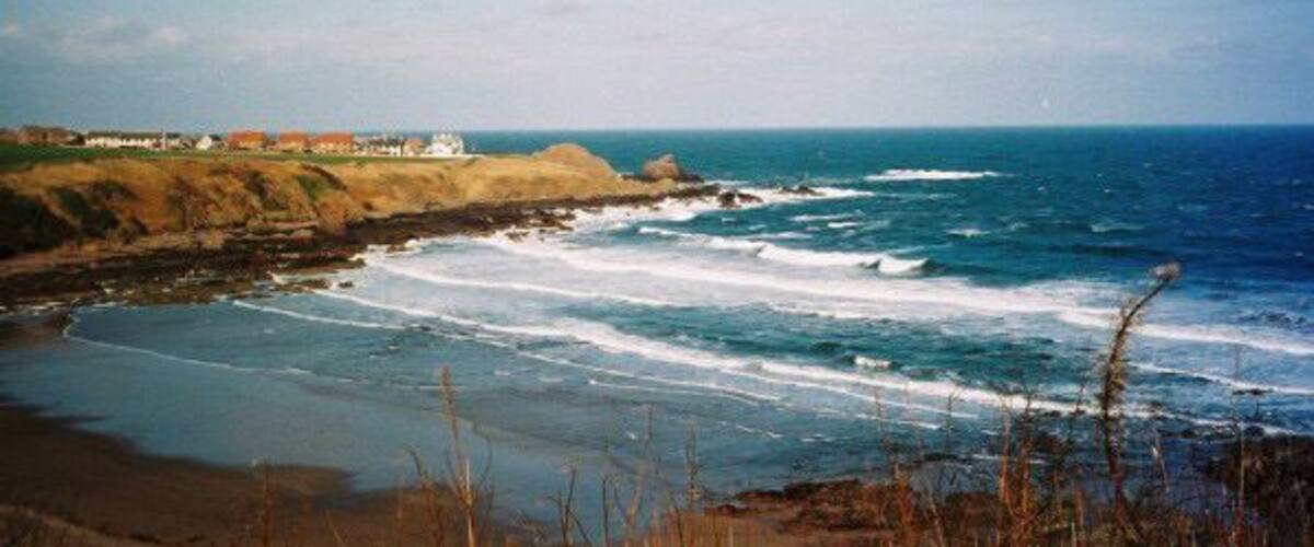 A coastal landscape with waves crashing onto the shore, a lighthouse, and a distant shoreline with buildings.