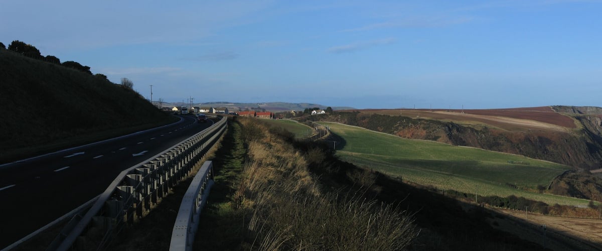 A coastal road with a guardrail, green fields, and cliffs overlooking the sea.