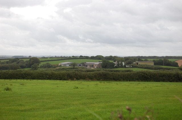 A rural landscape with green fields, a few buildings, and a cloudy sky.