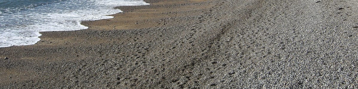 Ein Kiesstrand mit Wellen, Klippen und einer Person, die spazieren geht.