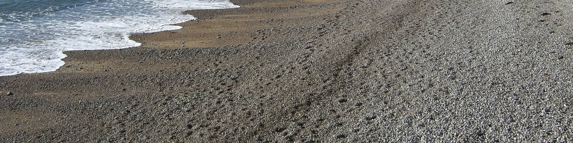 A pebble beach with waves, cliffs, and a person walking.