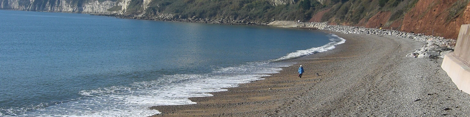 Una playa de guijarros con olas, acantilados y una persona caminando.