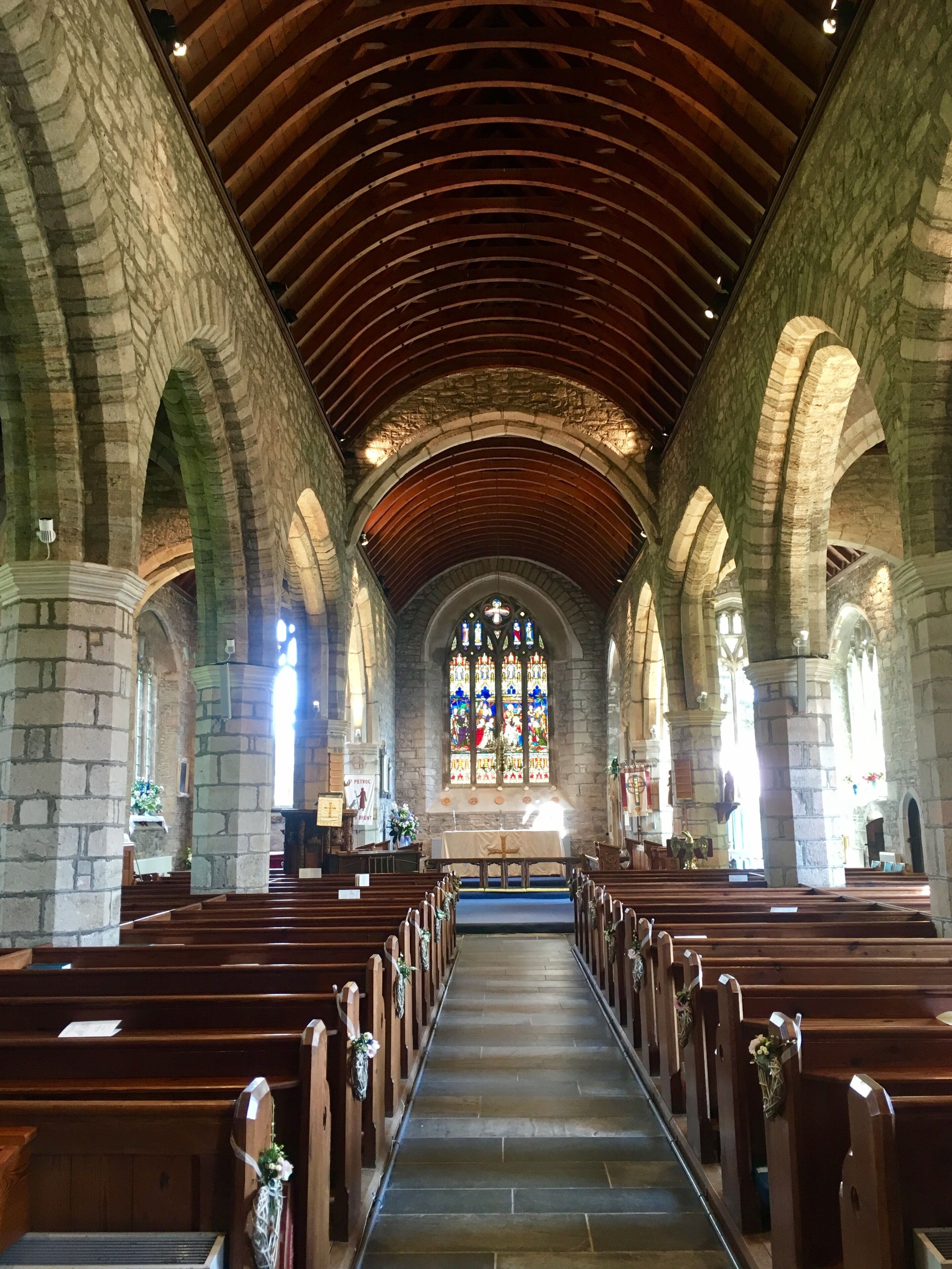 An interior of a church with stone arches, wooden pews, and a stained glass window.