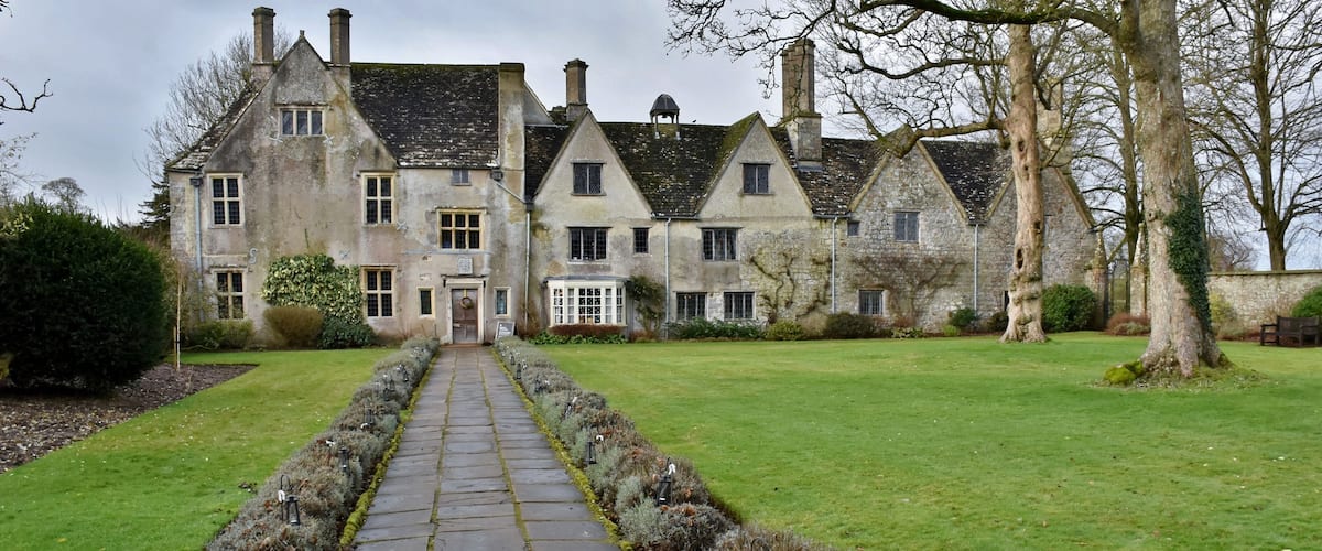A stone pathway leading to a large stone building with multiple chimneys and windows.