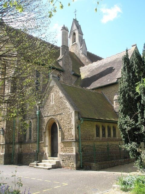A stone church with a steep roof, a bell tower, and a large tree in front.