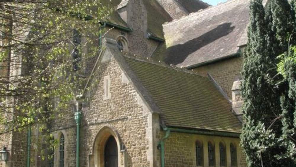 A stone church with a steep roof, a bell tower, and a large tree in front.
