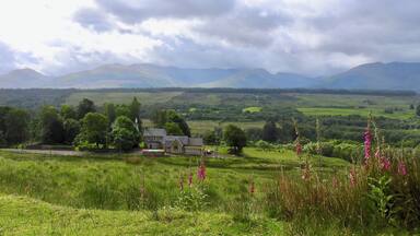 A rural landscape with a house, green fields, and mountains in the background.