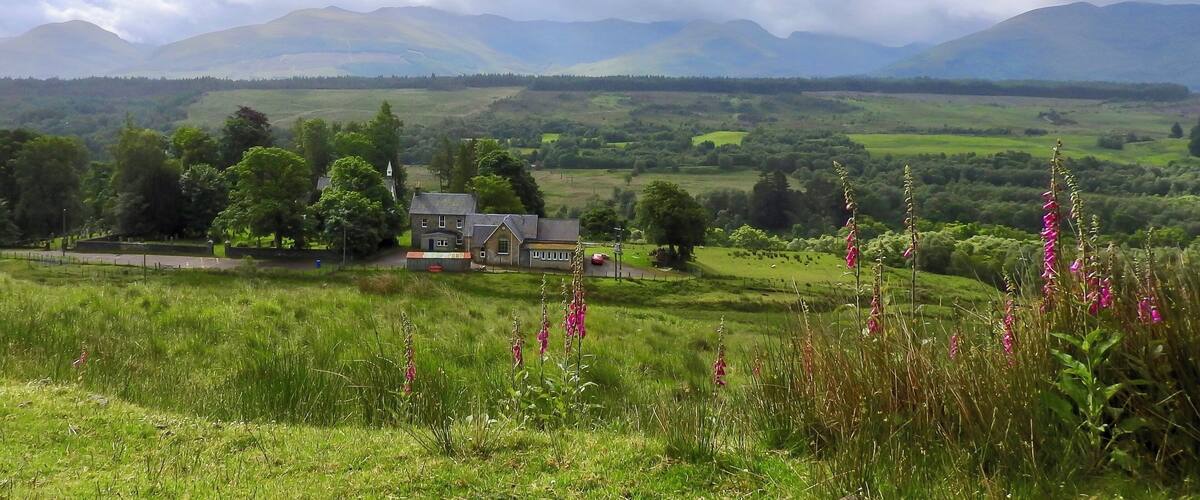 A rural landscape with a house, green fields, and mountains in the background.