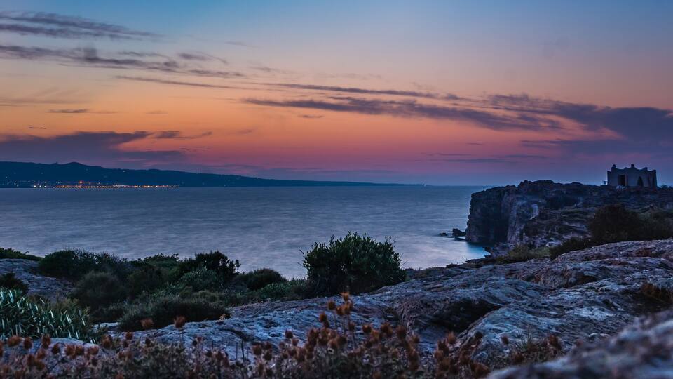 A coastal landscape with a rocky outcrop and a small building, likely a lighthouse, overlooking the sea.
