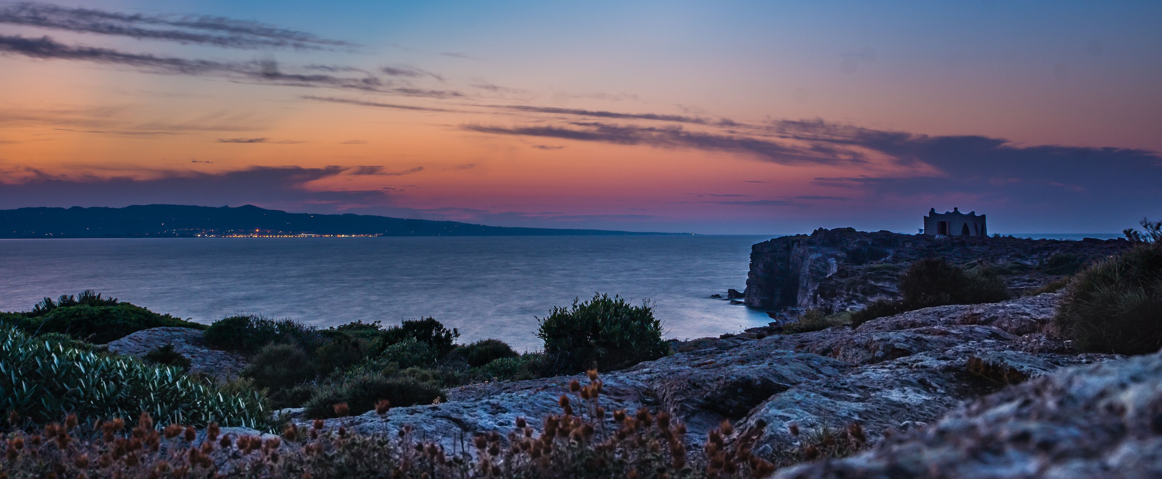 A coastal landscape with a rocky outcrop and a small building, likely a lighthouse, overlooking the sea.