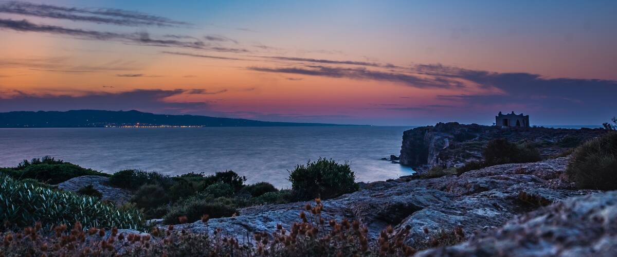 A coastal landscape with a rocky outcrop and a small building, likely a lighthouse, overlooking the sea.