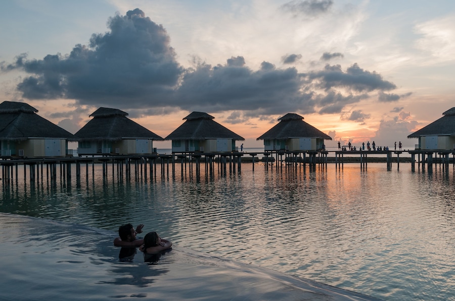 Ein Sonnenuntergang über einer Reihe von Bungalows, die auf Stelzen über dem Wasser gebaut sind, mit einem Paar im Vordergrund.