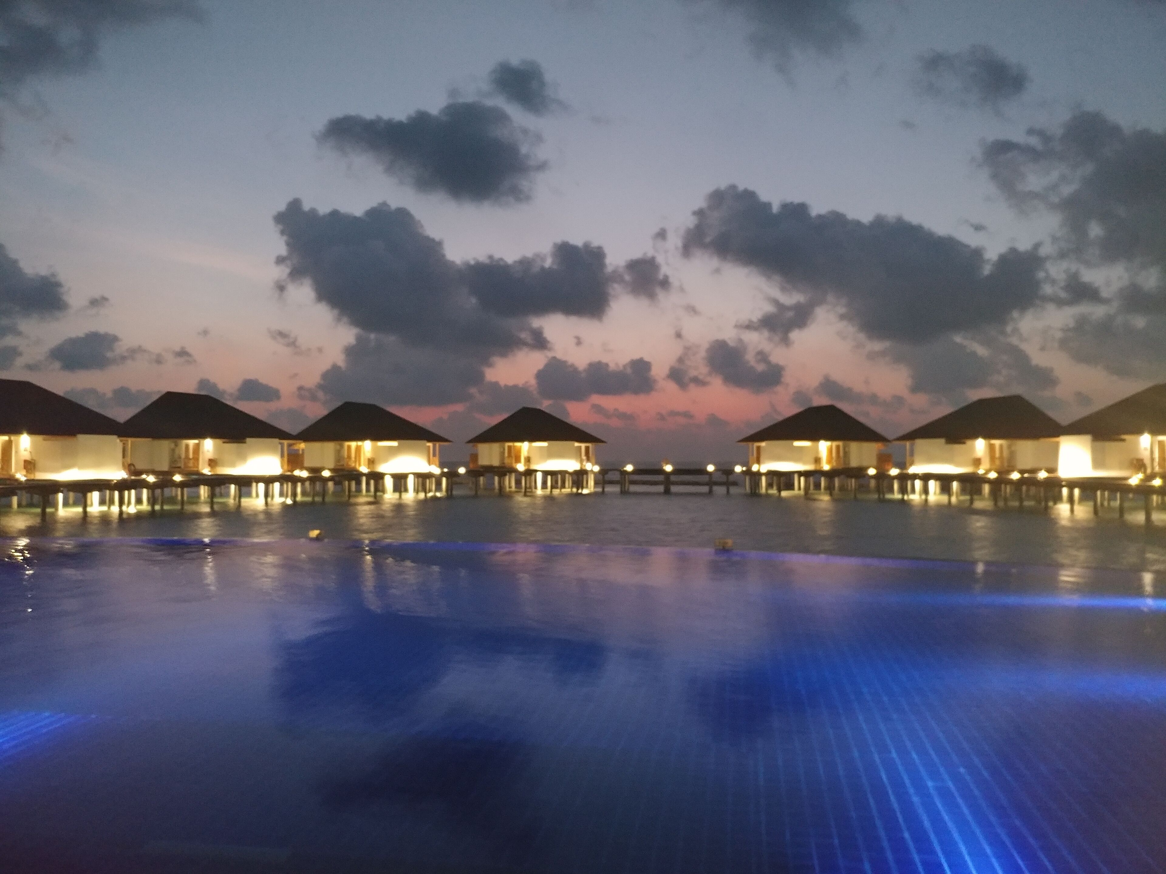 A series of overwater bungalows with illuminated windows and a calm pool reflecting the sky.