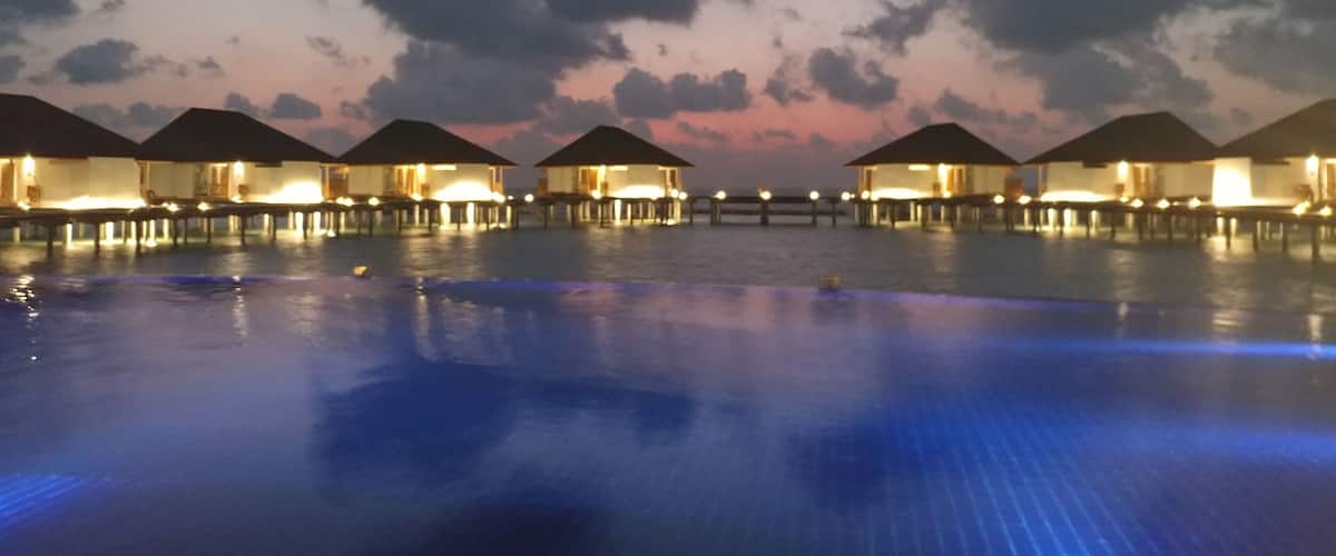 A series of overwater bungalows with illuminated windows and a calm pool reflecting the sky.