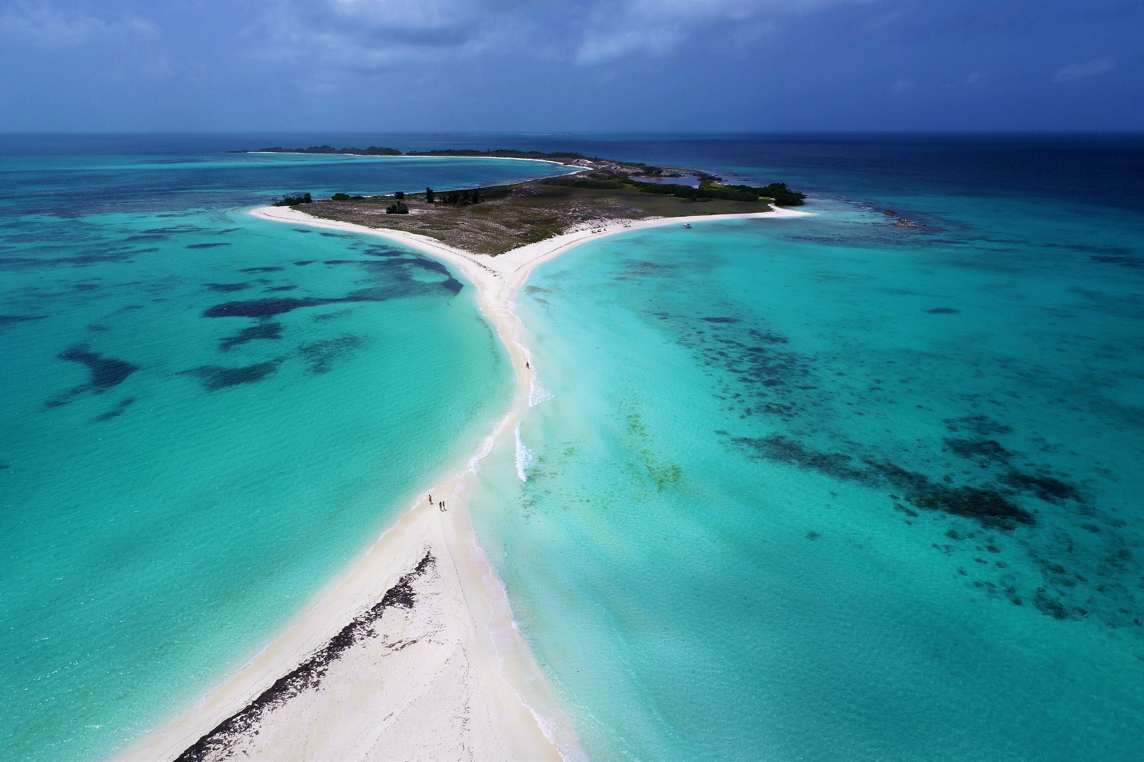 An aerial view of a turquoise beach with a white sand strip leading to a small island with ruins.