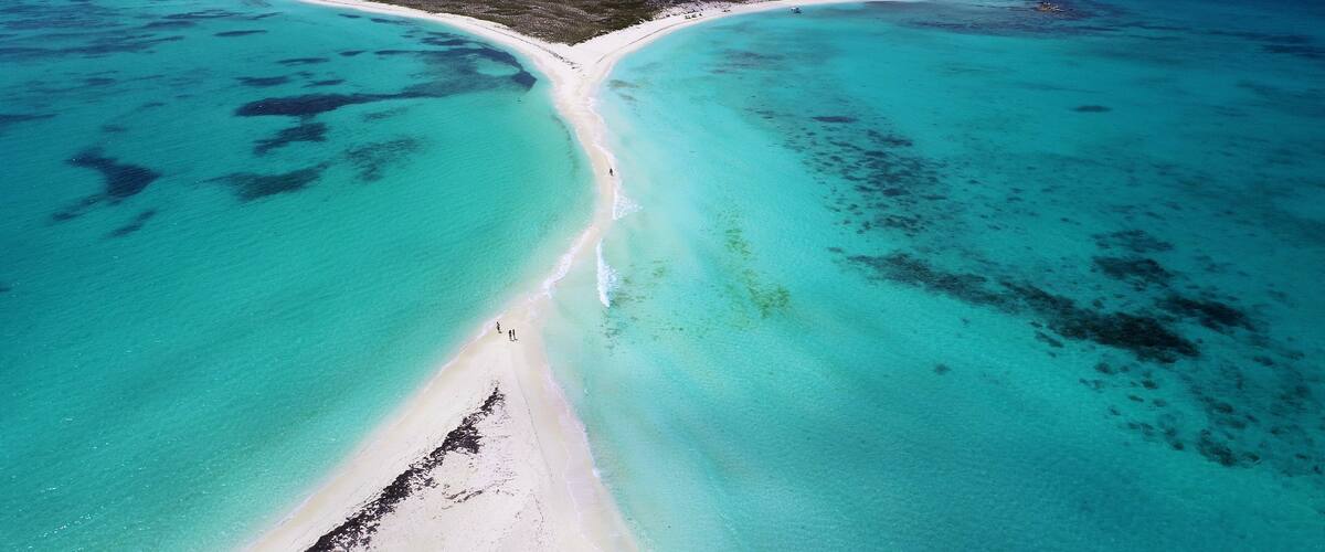 An aerial view of a turquoise beach with a white sand strip leading to a small island with ruins.