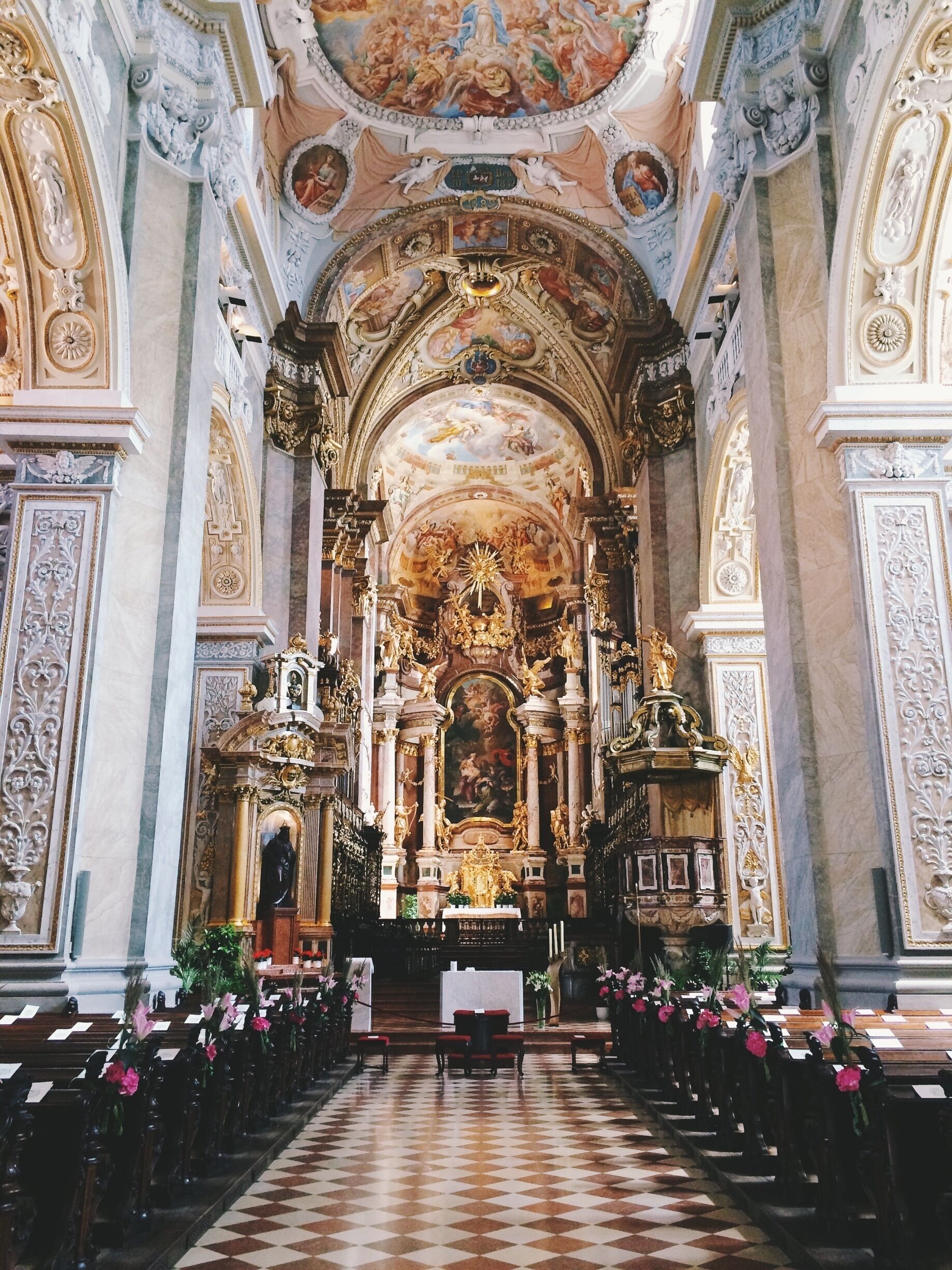 An ornate church interior with a checkered floor, rows of pews, and elaborate ceiling frescoes.