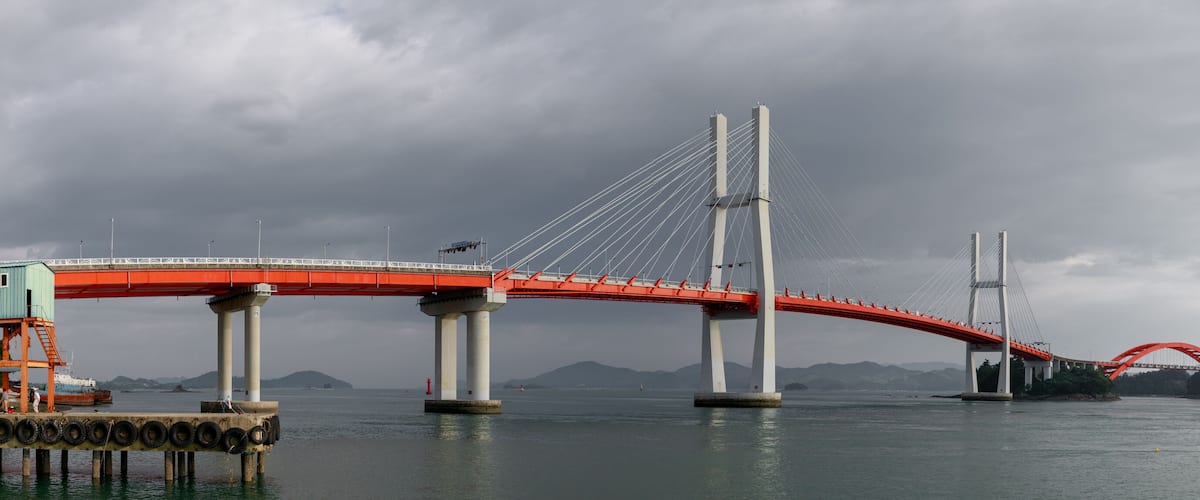 A large suspension bridge with red railings and cables, spanning a body of water with mountains in the background.