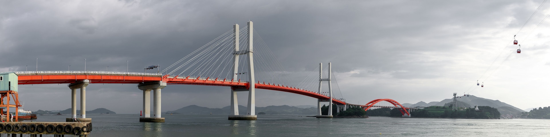 A large suspension bridge with red railings and cables, spanning a body of water with mountains in the background.