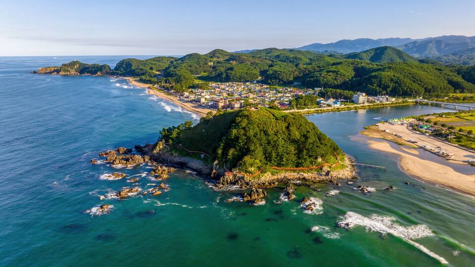 An aerial view of a coastal town with a prominent hill, clear blue waters, and a bridge connecting the mainland to an island.