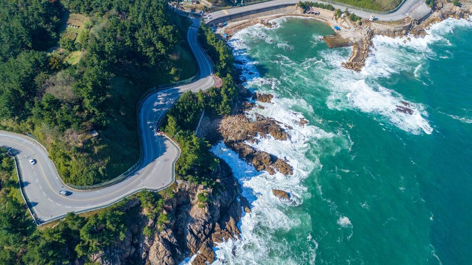 An aerial view of a coastal road winding through rocky cliffs and dense forests, with waves crashing against the shore.