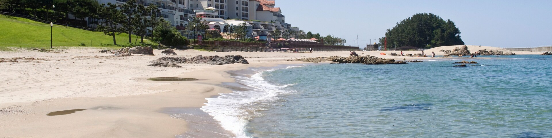 Uma praia com águas cristalinas, areal arenoso e um grande edifício de resort com telhados vermelhos.