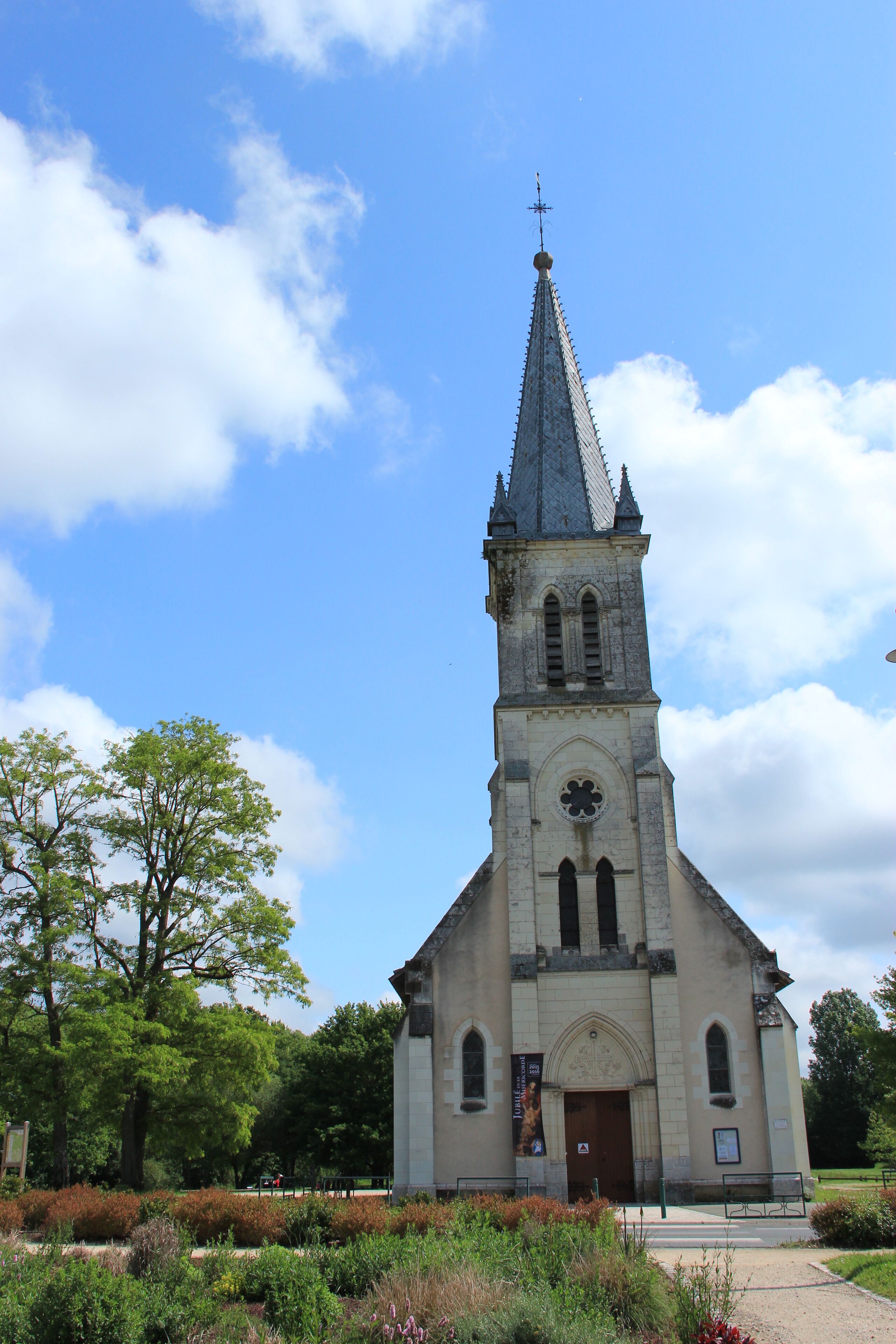 A church with a tall spire and a cross at the top, surrounded by greenery and trees.