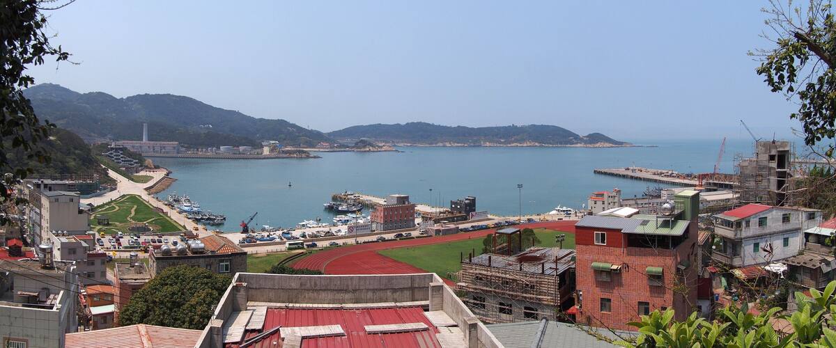 A coastal cityscape with a marina, a running track, and mountainous terrain in the background.