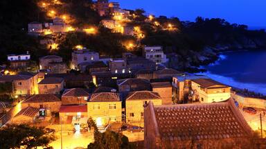 A coastal village at dusk with illuminated buildings and a steep hillside.
