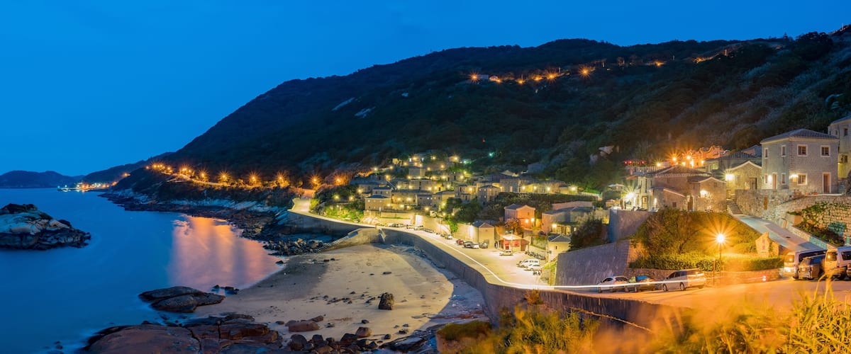 A coastal town at dusk with illuminated buildings, a beach, and a rocky shoreline.