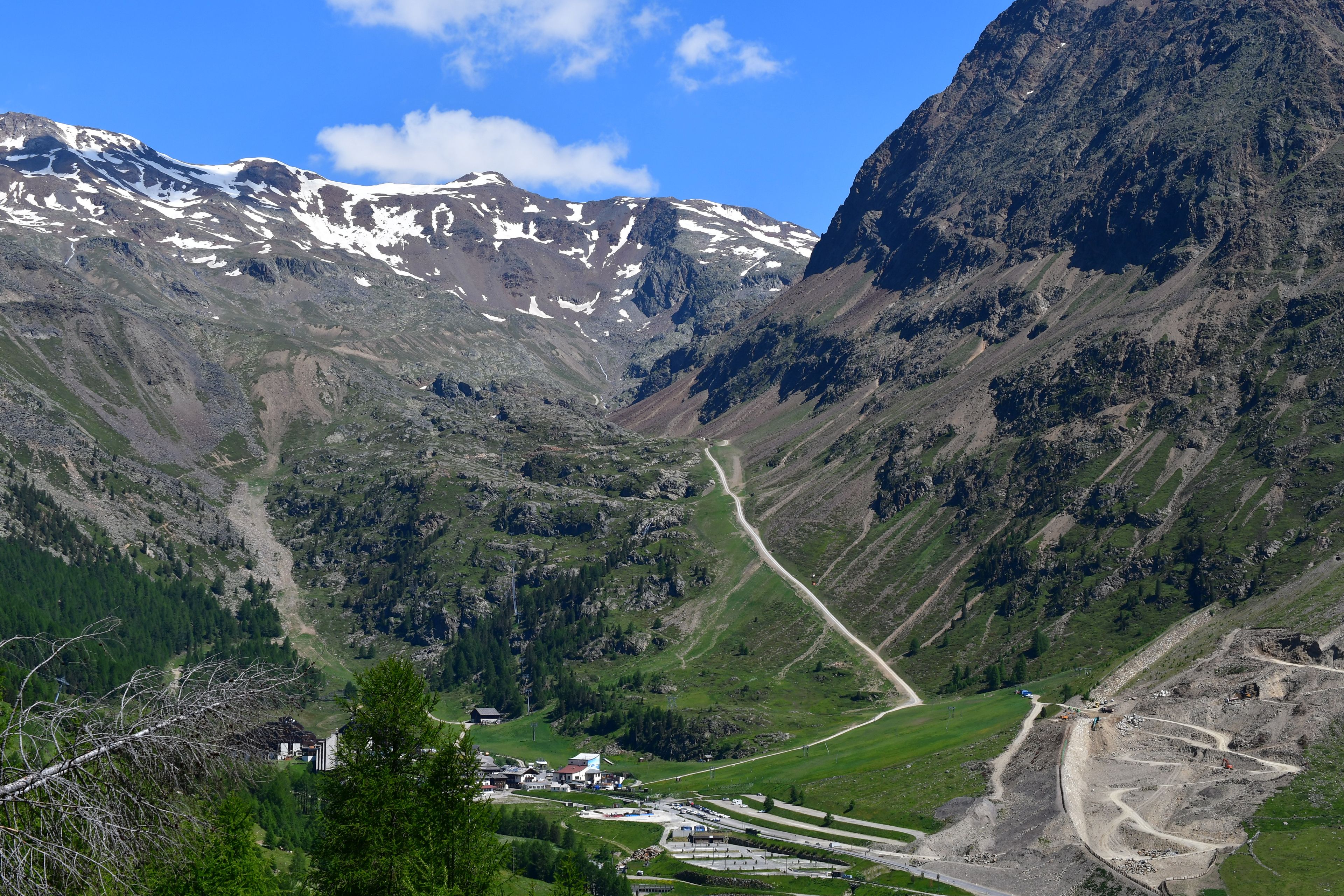 A mountainous landscape with a road winding through a valley, a small settlement with buildings, and snow-capped peaks in the background.