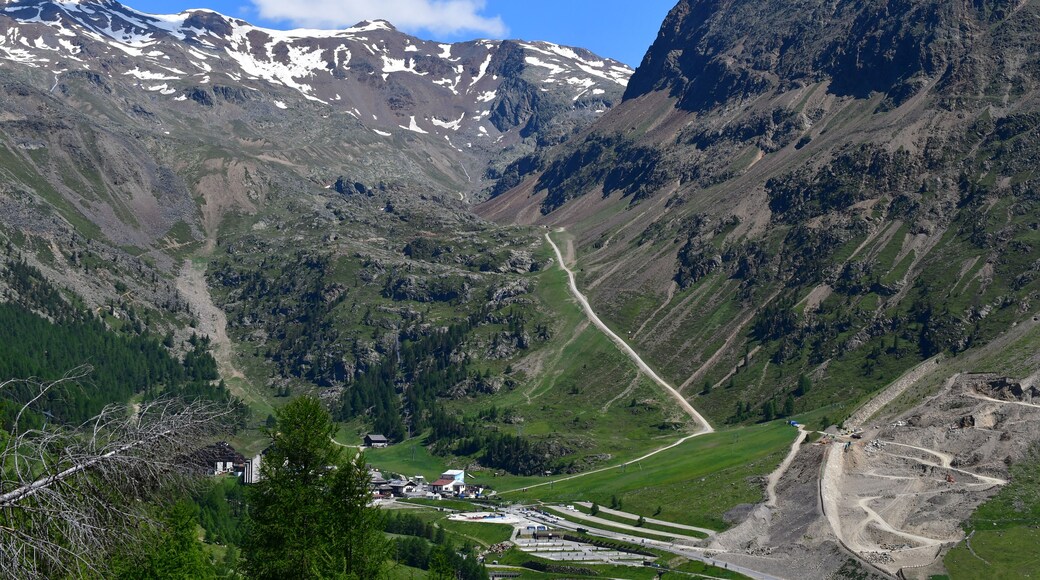 A mountainous landscape with a road winding through a valley, a small settlement with buildings, and snow-capped peaks in the background.