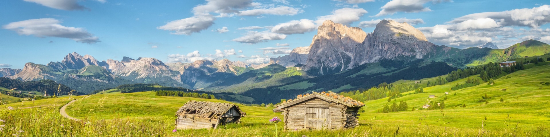 Eine Berglandschaft mit zwei rustikalen Holzhäusern, einem gewundenen Pfad und blauem Himmel.