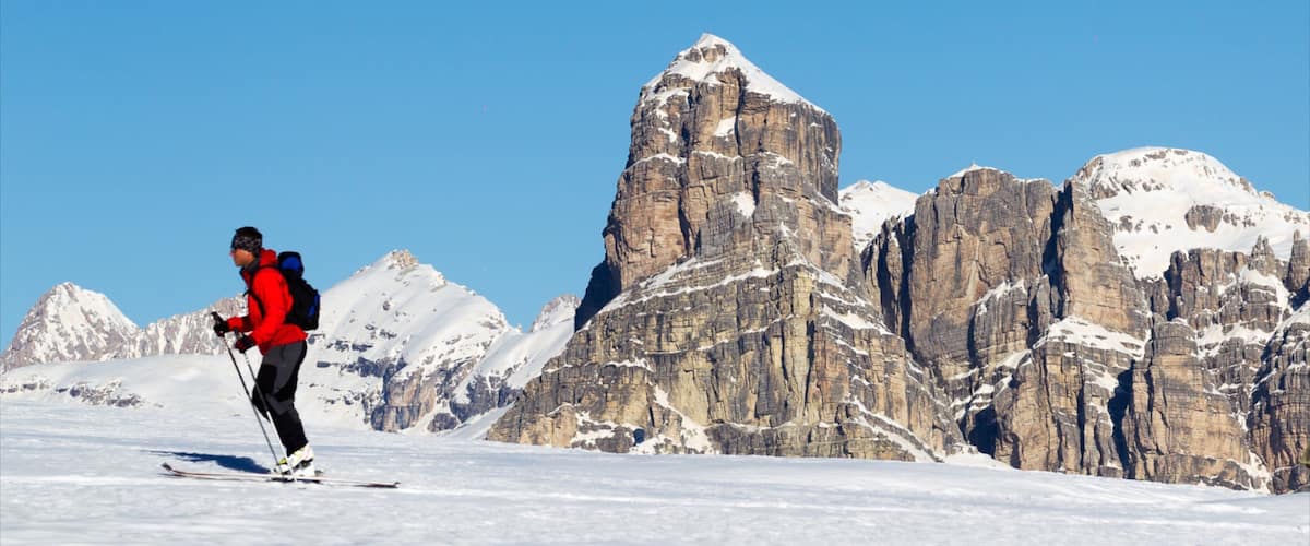 A skier in a red jacket on a snowy mountain with rocky cliffs in the background.