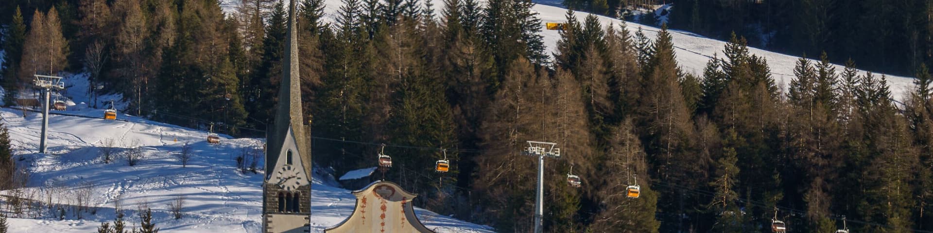 A snow-covered mountain town with a church, houses, and a ski lift against a backdrop of rugged mountains.