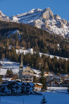 Un pueblo cubierto de nieve con una iglesia, casas y un telesilla frente a montañas escarpadas.