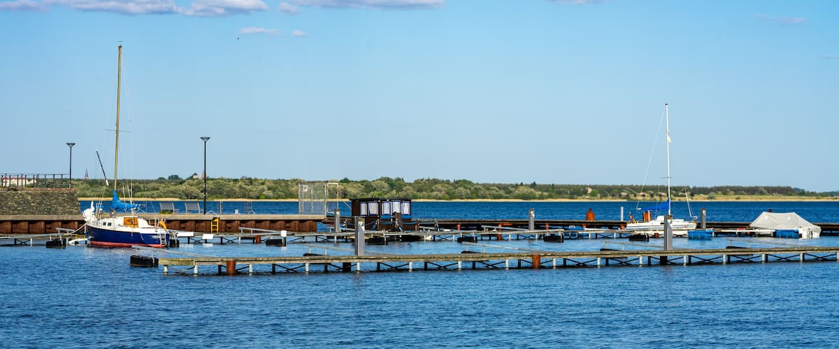 A harbor with boats docked at piers, a lighthouse, and a distant hill with vegetation.