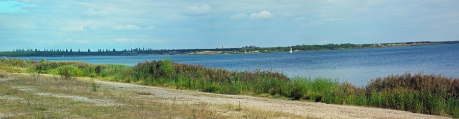 A river with boats, a distant shoreline with trees and buildings, and a dirt path along the riverbank.