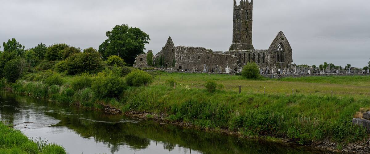 A historic stone castle with a tall tower, surrounded by lush greenery and a calm river.