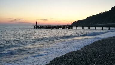 A pebbled beach with waves at sunset, a pier extending into the water, and a hill with trees in the background.